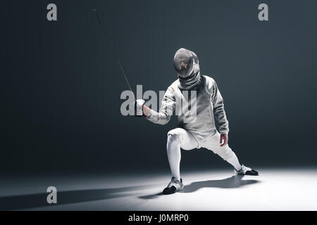 Tireur professionnel dans l'escrime masque avec rapière en position debout sur gris Banque D'Images