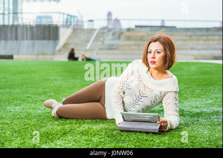 American College student studying in New York, le port de chandail en tricot blanc, marron pantalon long, bottes, couché sur pelouse verte sur le campus, la lecture en comprimé comp Banque D'Images