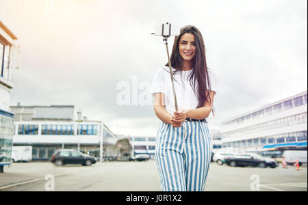 Vue grand angle du souriant belle femme prenant sa propre photo avec l'appareil photo sur stick selfies au milieu de rue sous ciel nuageux avec copie savs Banque D'Images