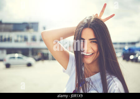 Fun jeune femme faisant un geste cornes derrière sa tête avec ses doigts comme elle sourit à l'appareil photo à l'extérieur de la ville Banque D'Images
