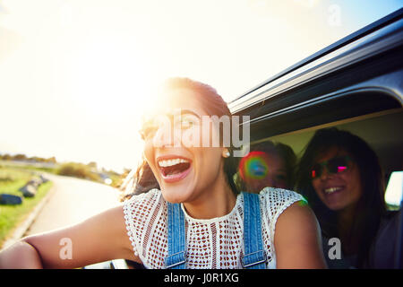 Happy young woman leaning excité sortir d'une voiture rétro de rire par une vive poussée du soleil matinal comme elle et ses amis vont sur un voyage sur la route ensemble Banque D'Images