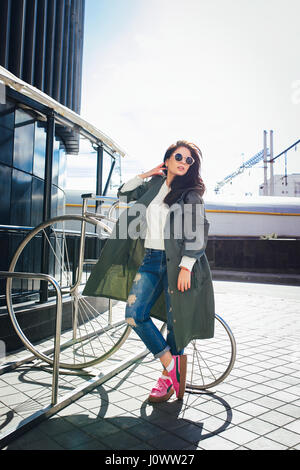 Fashion closeup portrait de nice jolie jeune femme posant en lunettes de hipster .de plein air heureux Brunette girl in green rose imperméable et des chaussures de marche de la rue de la ville. Banque D'Images