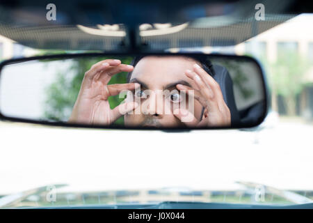 Closeup portrait drôles, jeune homme à la recherche du pilote à l'arrière, rétroviseur intérieur en essayant de rester éveillé à l'aide de mains pour ouvrir les yeux, l'intérieur du pare-brise de voiture b isolés Banque D'Images