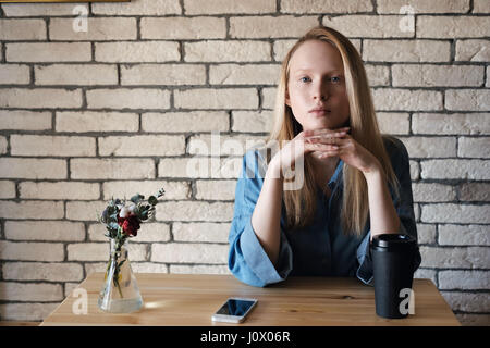 Une jeune blonde dans une chemise bleue, est assis à une table dans un café, pose sa tête sur ses mains. Sur la table il y a une tasse de café avec du papier noir et Banque D'Images