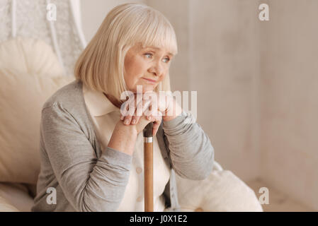 Tristes pensées. Déprimé malheureux vieille femme appuyée sur son bâton de marche et d'être impliqué dans les pensées tout en restant assis dans le fauteuil Banque D'Images