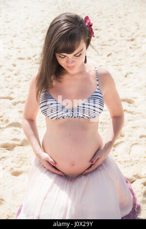 Pregnant woman sitting on beach, berçant son ventre Banque D'Images