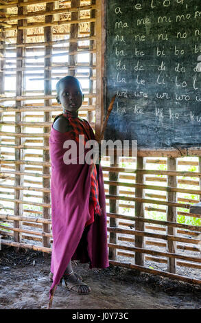 Jeune élève de Maasai souriant devant le tableau noir à l'école du village apprenant des voyelles anglaises vêtues d'un Shuka rouge traditionnel. Banque D'Images