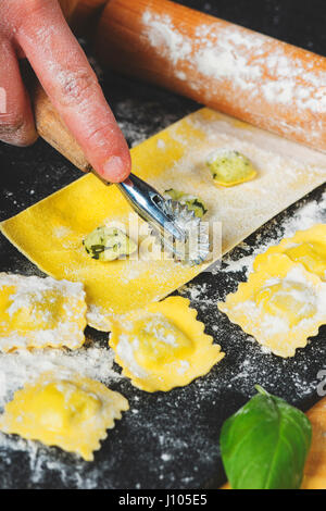 Préparation de raviolis dans la cuisine avec des outils et ingrédients : pâte, farine, œufs, farce, couteau, rouleau, planche. Découpe des carrés pour terminer la préparation. Banque D'Images