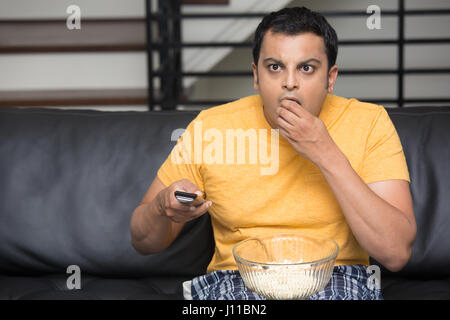 Closeup portrait, jeune homme en t-shirt jaune, assis sur le canapé de cuir noir, regarder la télévision, holding remote, surpris de ce qu'il voit, mâchonnant popcor Banque D'Images