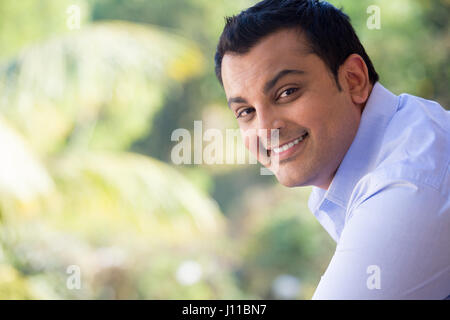 Closeup portrait of handsome young man in blue shirt debout à l'extérieur sur son balcon, isolé à l'extérieur l'extérieur arrière-plan avec des arbres verts Banque D'Images