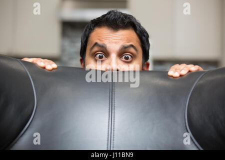 Closeup portrait jeune homme cultivé, se cacher derrière un canapé en cuir noir, de peur et d'inquiétude, isolé à l'intérieur arrière-plan Banque D'Images