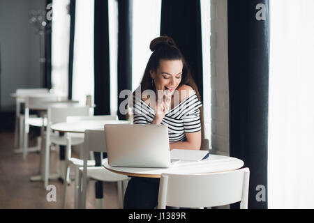 Photo de jolie femme en jouant avec un stylo et la réflexion sur un sujet tout en restant assis dans un café shop avec son ordinateur portable. Banque D'Images