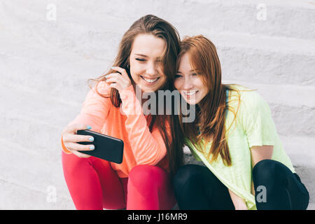 Deux belles jeunes filles assis sur les escaliers, à la recherche de téléphone intelligent et de rire. Les amies se reposer à l'été. Banque D'Images