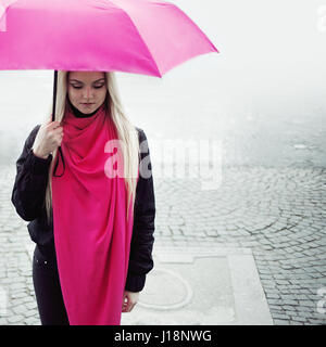 Triste pensive Girl standing sous un parapluie lumineux sur la rue. La notion de positivité et d'optimisme. Fille dans une écharpe et parapluie rose vif marcher dans une ville pluvieuse. Banque D'Images