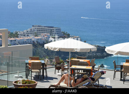 Blonde mature femme en maillot de bain qui est en train de lire couché dans l'ombre sous un parasol sur l'espace extérieur cafe à Los Gigantes resort sur l'eau de l'océan bleu backgrou Banque D'Images