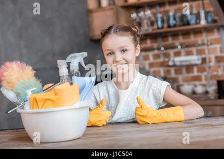 Adorable petite fille avec produits de nettoyage showing Thumbs up à la maison Banque D'Images