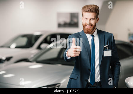 Vendeur in suit showing thumb up et à la caméra au concessionnaire en beauté Banque D'Images