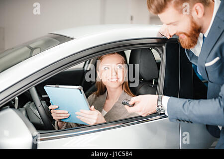 Vendeur barbu donnant la clé de voiture young woman sitting in new car with digital tablet Banque D'Images