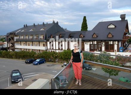 Hauts smiling blonde woman in red Pantalon et chemisier noir est debout sur la passerelle au-dessus de la route au matin nuageux sur fond d'hôtel Santa Susa Banque D'Images