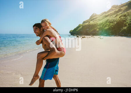 Vue de côté tourné de beau jeune homme s'occuper sa petite amie sur le dos à la mer. Man piggybacking woman on the beach. Banque D'Images