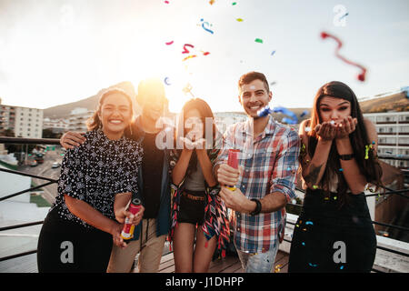 Groupe d'amis ensemble et soufflant les confettis sur rooftop party. Banque D'Images