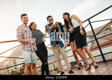 Jeune homme de l'ouverture d'une bouteille de champagne avec des amis. Les jeunes fête avec champagne au travail sur le toit. Banque D'Images