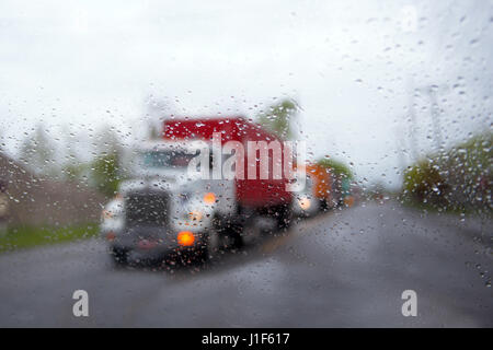 Le convoi chargé gros camion semi camions avec remorques et conteneurs sur la route par temps de pluie à travers le pare-brise avec des gouttes de pluie. Banque D'Images