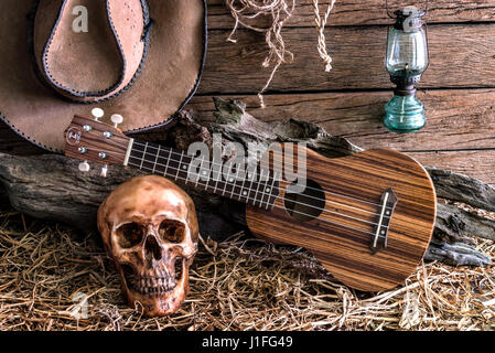 Still Life crâne humain et ukulele sur la paille avec des bottes en cuir et de l'ouest américain Rodeo Cowboy hat feutre brun, fond sombre et vintage à Banque D'Images