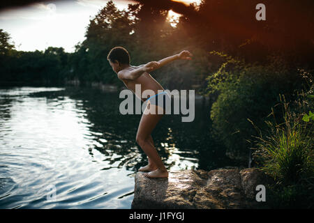 Garçon sautant de la falaise dans un lac au coucher du soleil Banque D'Images