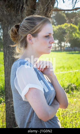 Portrait of young blonde woman à l'avant et à l'écart de l'article appareil photo à l'ombre sous un arbre dans un cadre rural - photo verticale Banque D'Images