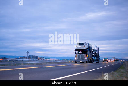 Big Rig semi truck avec une remorque pour véhicule de transport avec des voitures à plusieurs niveaux sur la route le soir de l'avant avec les phares de voiture de convoi Banque D'Images