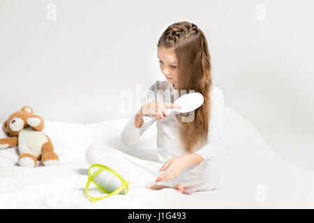 Adorable petite fille en pyjama assis sur le lit et peigner les cheveux Banque D'Images