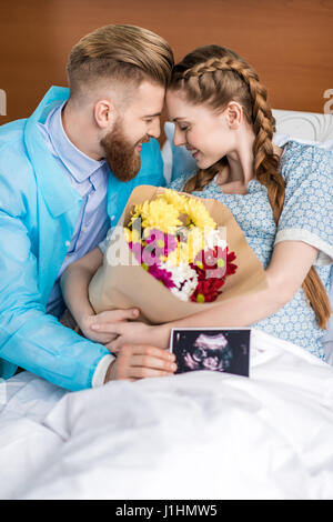 Femme enceinte avec mari hugging and holding sonogram et fleurs à l'hôpital Banque D'Images