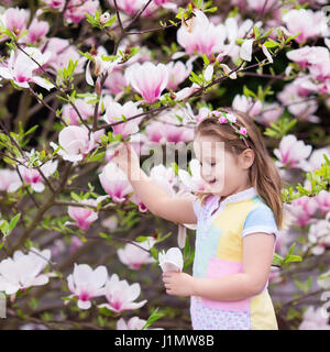 Happy little girl de couleur pastel robe et couronne de fleurs rose jouer sous magnolia grandes fleurs cueillette dans le jardin de printemps en fleurs. Enfant avec ma Banque D'Images