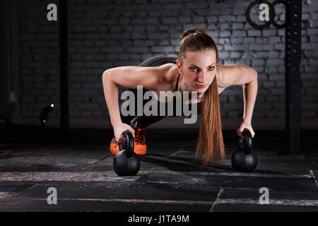 Belle jeune femme sportive dans le sportwear noir faisant un pompes avec kettlebell contre mur de brique à la salle de sport crossfit. Close-up photo horizontale. Banque D'Images