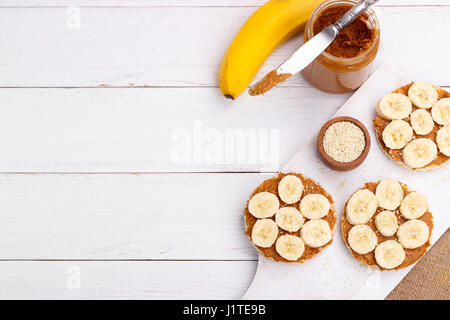 Gâteaux de riz avec du beurre d'arachide et de tranches de banane et graines de sésame sur table en bois blanc Banque D'Images