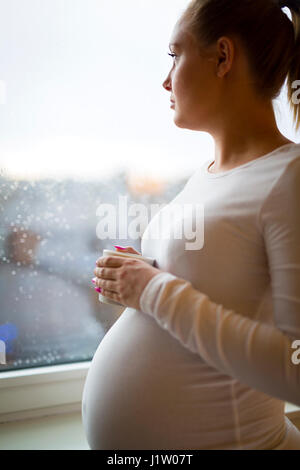 Une femme enceinte en regardant par la fenêtre plateau drinkng Banque D'Images