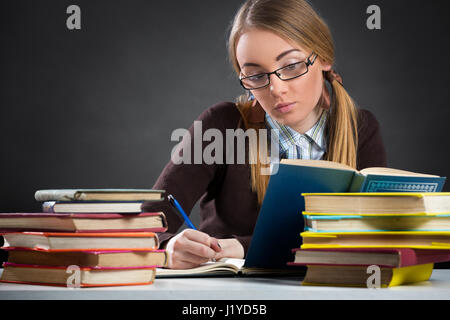 Fille étudiante à lunettes assis à un bureau rempli de livres et de travail à domicile Banque D'Images