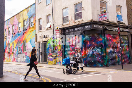 Couple Hipster traversent la route en tirant un landau dans Redchurch street où il rencontre Chance Street. Les bâtiments couverts de graffitis et de peintures murales à l'arrière Banque D'Images