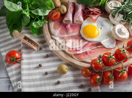 Petit-déjeuner méditerranéen sain d'épicerie : œufs frits, jambon, champignons, tomates, olives, basilic, romarin sur plateau en bois Banque D'Images