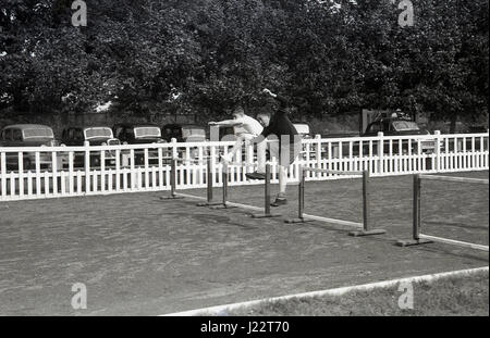 Dans les années 1950, historique, en dehors d'une piste de course à pied de cinder, deux jeunes garçons en compétition dans une course à obstacles franchissent des barrières ou des obstacles, avec des voitures de l'époque garées sur une ligne derrière une clôture blanche, en Angleterre, au Royaume-Uni. Banque D'Images