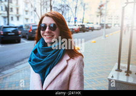 Belle jeune fille aux cheveux rouge est à pied par la rue dans une robe rose et bleu foulard, avec des lunettes. Banque D'Images