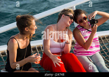 Groupe de touristes masculins et féminins ayant un verre alors que sur une croisière en catamaran au coucher du soleil au large de la côte d'Aruba, des Caraïbes. Banque D'Images