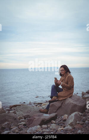 Woman using mobile phone while sitting on rock at beach against sky Banque D'Images