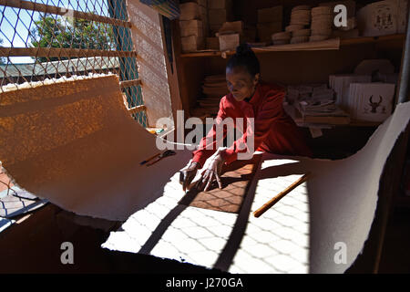 Coupe d'une femme d'une feuille de papier fait main à l'usine de fabrication de papier à Ambalavao Madagascar ; les produits de papier fini sur une étagère derrière elle Banque D'Images