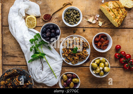 Tapenade d'olive dans une assiette blanche photographié en vue de dessus. Trois types d'olives, câpres, tomates séchées, herbes de tranches de pain de maïs l'accompagnent. Banque D'Images