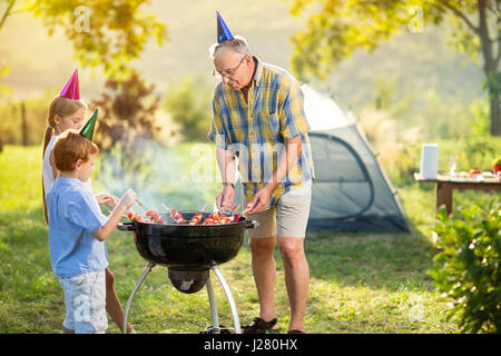 Les enfants et grand-père ayant une partie de barbecue sur camping Banque D'Images