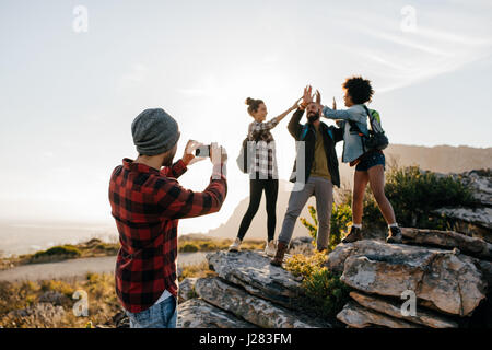 Jeune homme à prendre des photos de ses amis faisant cinq haut en campagne tout en randonnée. Groupe de randonneurs bénéficiant pendant les vacances d'été. Banque D'Images