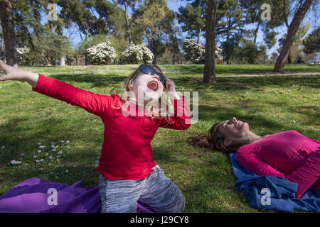 Trois ans, l'enfant blond avec chemise rouge, assis sur les genoux dans l'herbe verte dans le parc, à côté de mère dormir, avec de grandes lunettes de soleil femme adultes singin Banque D'Images