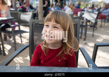 Portrait d'enfant de trois ans, blonde, visage avec chemise rouge, drôle de rire, avec des miettes dans la bouche, assis à l'extérieur terrasse bar café avec ta gris Banque D'Images